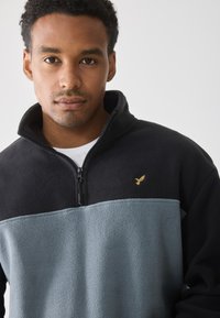 Sudadera de forro polar en negro y azul claro, con un cuello alto, cierre de cremallera y un acento de logo dorado en el pecho. Textura suave.
