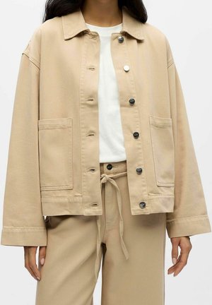 Femme portant une veste beige ample boutonnée sur une chemise blanche avec un pantalon beige à cordons assorti, debout devant un fond uni.