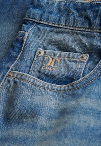 Nærbillede af lomme på blå denimjeans med metalknapper og et lille syet mærke med teksten "CULT" under en dobbelt-C-logo.