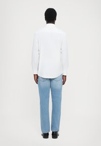 Hvit langermet skjorte med buet kant, kombinert med lyseblå straight-leg jeans og svarte sko, sett bakfra.