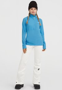 Ljusblå långärmad zip-up-tröja med hög krage och vita snöbyxor. Svart mössa synlig. Byxorna har en logotyplapp.