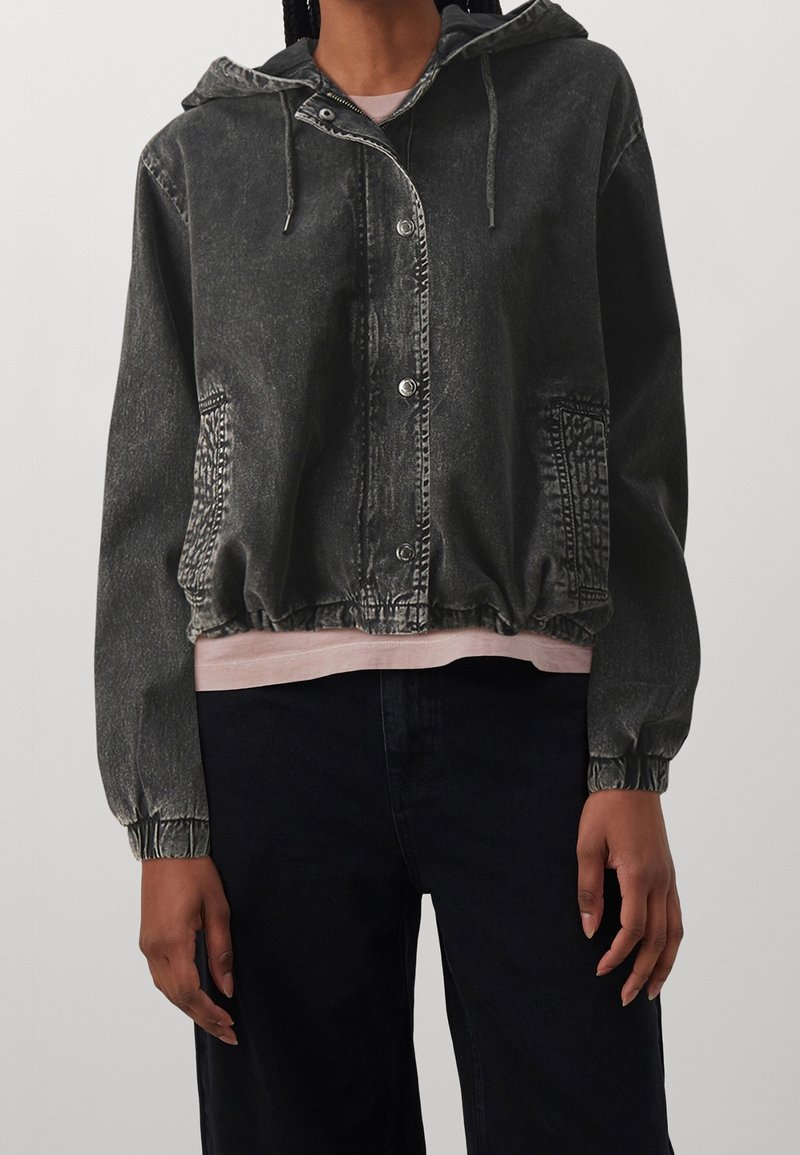 Personne portant une veste noire à capuche délavée avec boutons-pression, poignets élastiques et poches avant par-dessus une chemise rose clair et un pantalon noir.