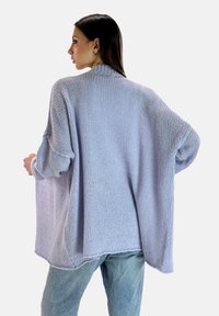 Cardigan de punto lavanda con un corte holgado, mangas anchas y cuello acanalado. Textura suave y diseño de espalda abierta. Combinado con jeans de color azul claro.