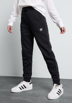 Osoba nosząca czarne spodnie dresowe Adidas oraz białe buty sportowe Adidas stojąca na szarej podłodze na jasnym tle.