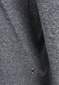 Grå stickad tröja med ett textuerat mönster. Har en ribbad nederkant och en liten logotyp patch på vänster sida längst ner. Mjukt tygutseende.