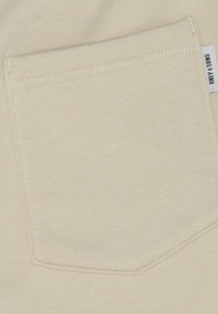 Beige stof med en vævet struktur, prydet med en rektangulær påsyet lomme med synlige sømme og et mærke på den sidekant.