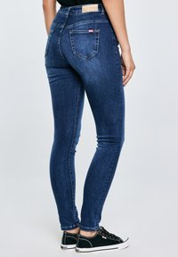 Persoană purtând blugi skinny albastru închis cu buzunare în spate și adidași negri cu talpă joasă, stând pe un fundal simplu, luminos.