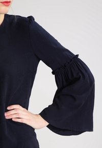Blusa navy con maniche lunghe a campana, caratterizzate da volant arricciati e una sottile trama floreale. Materiale liscio e corpetto aderente.
