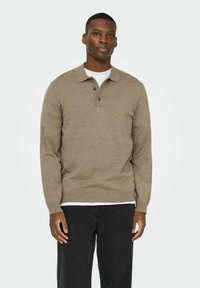 Jersey tejido de color marrón claro con cuello con solapa, tres botones, mangas largas y dobladillo acanalado, llevado sobre una camisa blanca.