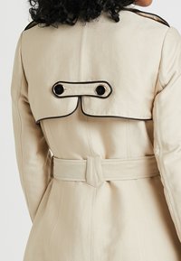 Beżowy trencz z usztywnionym tyłem, czarnym obszyciem i podwójnymi czarnymi guzikami. Posiada pasek w talii dla dopasowanego wyglądu.