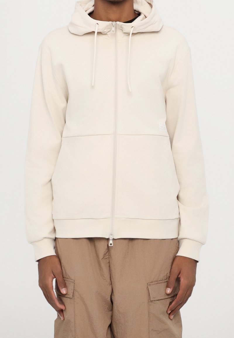Personne portant un sweat à capuche crème zippé avec capuche et cordons, associé à un pantalon cargo marron clair avec poches avant.
