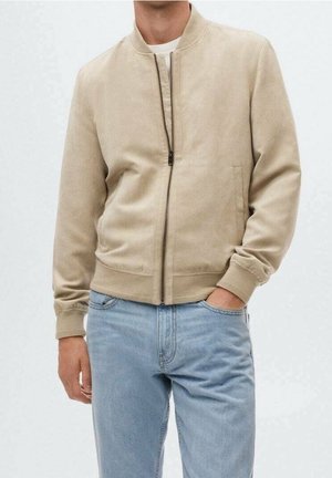 Mand iført beige bomberjakke med lynlås og ribkantede ærmer samt lyseblå jeans, stående med den ene hånd i lommen.