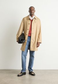 Beige jas met grote knopen, rode trui, lichtblauwe spijkerbroek en zwarte schoenen met een lichte accent. Houdt een zwarte tas vast, staande op een neutrale vloer.