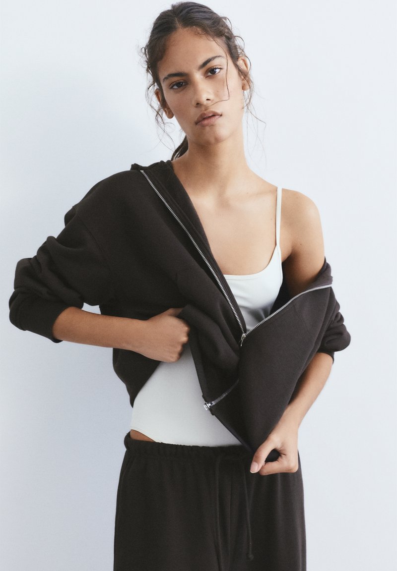 Fiatal nő fekete cipzáras dzsekit visel fehér trikó fölött, fekete nadrágban, egyszerű fehér háttér előtt.