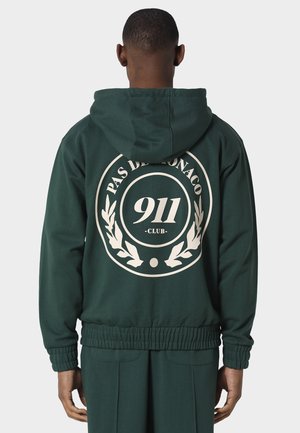 Man draagt een donkergroene hoodie en broek, achterkant gezien, met witte ronde "Pas De Monaco 911 Club"-logo op de hoodie.
