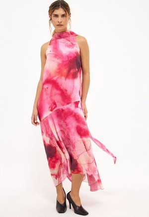 Naine kannab varrukateta roosat ja punast akvarellimustriga poolpikk kleiti asümmeetrilise äärega, mis on ühendatud mustade teravate ninaga kingadega.