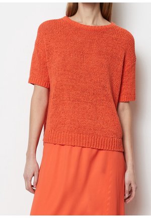 Kvinde iført en lys orange kortærmet strikket sweater og matchende orange nederdel, stående mod en ensfarvet hvid baggrund.