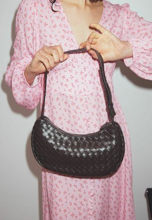 Austinti juodos odos rankinę su vingiuota siluetu, plonu petiniu diržu ir lygia tekstūra, laiko žmogus rožinės gėlių suknelėje.