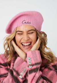 Rožinė megzta beretė su juodu užrašytu žodžiu "Crás". Modelis vilki derančią rožinę ir rudą dryžuotą paltą su sidabriniais mygtukais.