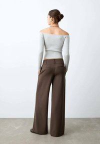Žena s drdolem nosí světle šedý žebrovaný top s odhalenými rameny a hnědé široké kalhoty, stojí na betonové podlaze s rukama v kapsách.