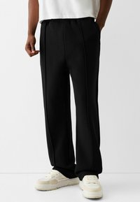 Black sweatpants with a relaxed fit, featuring two side pockets, vertical seams, and a drawstring waist. Paired with white sneakers.