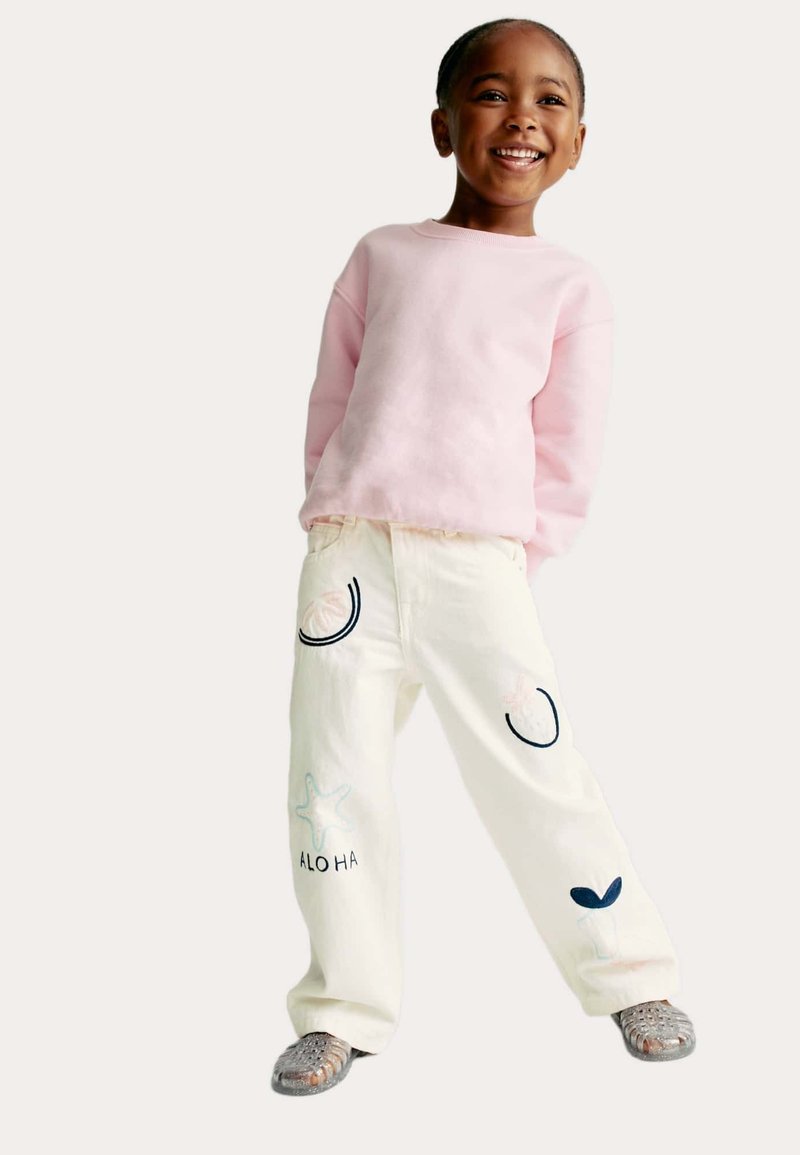 Sudadera rosa clara, pantalones blancos con acentos bordados coloridos (estrellas de mar, conchas y "ALO HA") y zapatos grises de texturizados sin cordones.