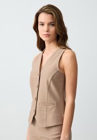 Gilet sans manches beige avec un décolleté en V profond, motif à rayures verticales, fermeture à boutons et une poche. Texture lisse et coupe ajustée.