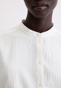 Biała koszula w paski z guzikami na przodzie i stójką, z perłowymi guzikami oraz gładką, lekką fakturą tkaniny.