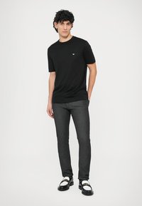 Svart t-shirt med korta ärmar och en liten vit logga på bröstet, kombinerad med mörkgrå slim fit-byxor och svartvita skor.