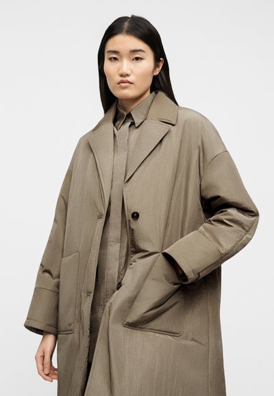 Νεαρή γυναίκα με ίσια σκούρα μαλλιά, φορώντας ένα φαρδύ παλτό ταουπέ και ένα ταιριαστό πουκάμισο, μπροστά σε απλό ανοιχτό φόντο.