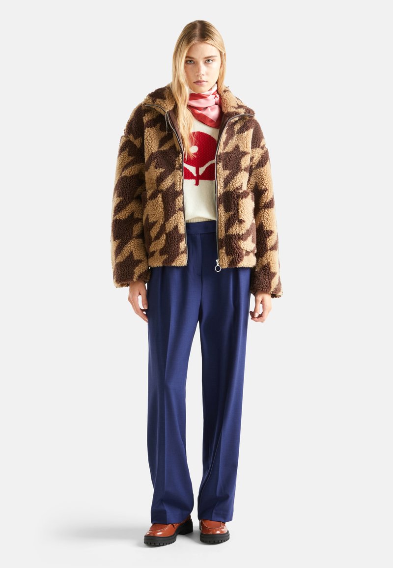 Braun-beige Hahnentritt-Fleecjacke, weißer Pullover mit rotem Grafikdruck, marineblaue weit geschnittene Hose und braune Plattformschuhe. Rosa Schal-Detail.