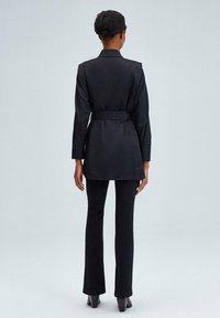 Blazer nero con cintura, dal design sartoriale, maniche lunghe e una texture liscia, abbinato a pantaloni neri aderenti e scarpe con tacco.