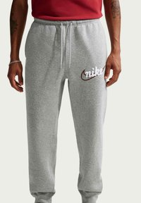 Bărbat purtând pantaloni sport Nike gri cu șnururi albe și tricou roșu, care arată tatuajele de pe încheietura mâinii și o brățară pe brațul stâng.