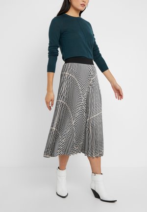 Femme portant un haut à manches longues vert foncé, une jupe midi plissée à carreaux noir et blanc, et des bottines blanches, debout sur un fond blanc uni.
