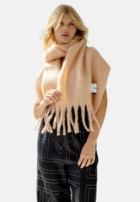 Écharpe douce de couleur pêche avec des bords frangés, fabriquée en matériau duveteux, enroulée autour des épaules, assortie à un pantalon noir à motifs.