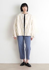 Quilted ivory jacket with diamond pattern, round neckline, large buttons; paired with light blue trousers and black loafers.