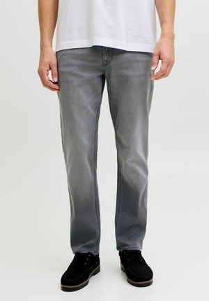 Mann trägt hellgraue Jeans mit geradem Schnitt, weißes T-Shirt und schwarze Schnürschuhe, steht vor einem einfarbigen hellen Hintergrund.