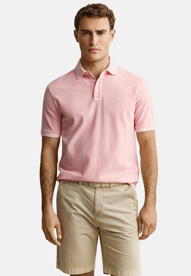 Homme portant un polo rose à manches courtes rentré dans un short beige avec ceinture, debout devant un fond blanc uni.