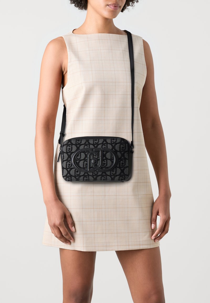 Borsa a tracolla nera con texture, caratterizzata da un design a pattern e un logo prominente. Indossata sopra un vestito a quadri chiari, con un profilo snello.