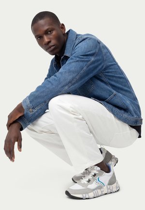 Uomo di colore accovacciato, con giacca di jeans blu, pantaloni bianchi e scarpe da ginnastica grigio-bianco con dettagli blu, su sfondo uniforme.