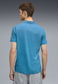 Turkizna polo majica s ovratnikom i kratkimi rokavi, ki ima gladko teksturo. Vidna je zadaj, vključuje nevsiljiv logo na rokavu.