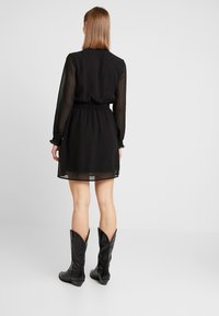 Schwarzes Kleid mit durchsichtigen langen Ärmeln, elastischem Bund und einem schwingenden Rock. Kombiniert mit knielangen schwarzen Stiefeln mit spitzen Kanten.