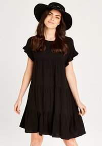Schwarzes, schichtweise gekleidetes Kleid aus weichem Stoff mit kurzen, gerafften Ärmeln und einer fließenden Silhouette. Kombiniert mit einem schwarzen Hut mit breiter Krempe.