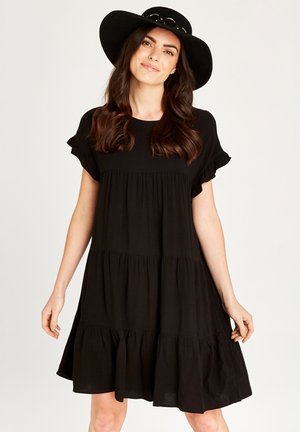Schwarzes, schichtweise gekleidetes Kleid aus weichem Stoff mit kurzen, gerafften Ärmeln und einer fließenden Silhouette. Kombiniert mit einem schwarzen Hut mit breiter Krempe.