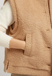 Gilet en laine beige avec une texture douce et duveteuse, doté de poches latérales, d'un col et d'une fermeture à boutons sur le devant. L'ourlet présente des fentes sur les côtés pour plus de confort.