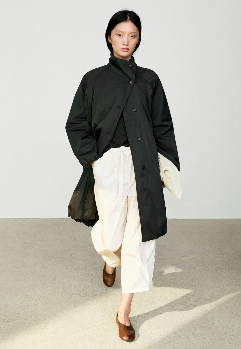 Woman walking indoors wearing a black buttoned coat, white wide pants, and brown slip-on shoes against a plain light background.