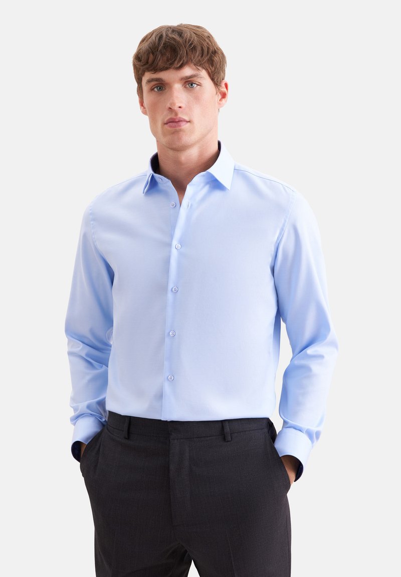 Homme portant une chemise bleu clair à manches longues rentrée dans un pantalon de costume noir, debout, les mains dans les poches.