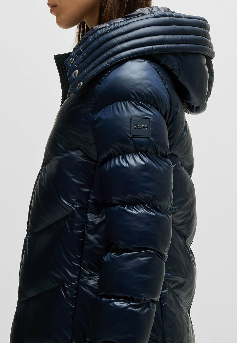 Granatowa kurtka puchowa z błyszczącym wykończeniem, pikowany wzór, oversize'owy ściągacz na kapturze oraz naszywka z logo na lewym rękawie. Zapięcie na zamek.