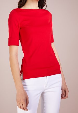 Vrouw draagt een aansluitend rood kortermig shirt met opgerolde manchetten, gecombineerd met een witte broek tegen een effen neutrale achtergrond.