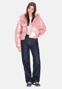 Błyszcząca różowa kurtka puchowa z kapturem, z zamkiem błyskawicznym i bocznymi kieszeniami, zestawiona z niebieskimi dżinsami i trampkami.